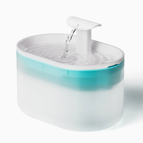 A modern oval pet water fountain with a translucent white body, a teal blue inner band, and a white pump dispenser pouring water into a clear tray, shown on a white background.